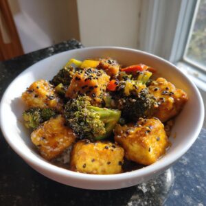Close-up of a white bowl filled with creamy almond butter tofu, broccoli florets, and bell peppers, sprinkled with sesame seeds.