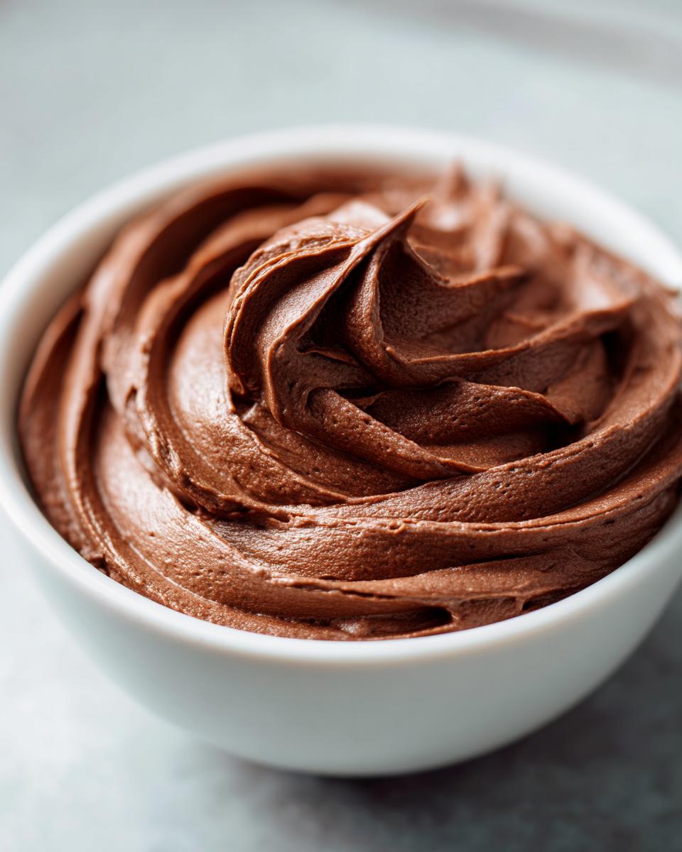 A close-up of smooth, rich chocolate frosting recipe swirled in a white bowl.