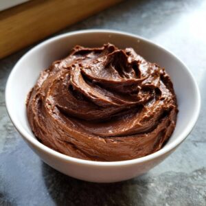 A close-up of rich, smooth chocolate fudge frosting swirled in a white bowl.
