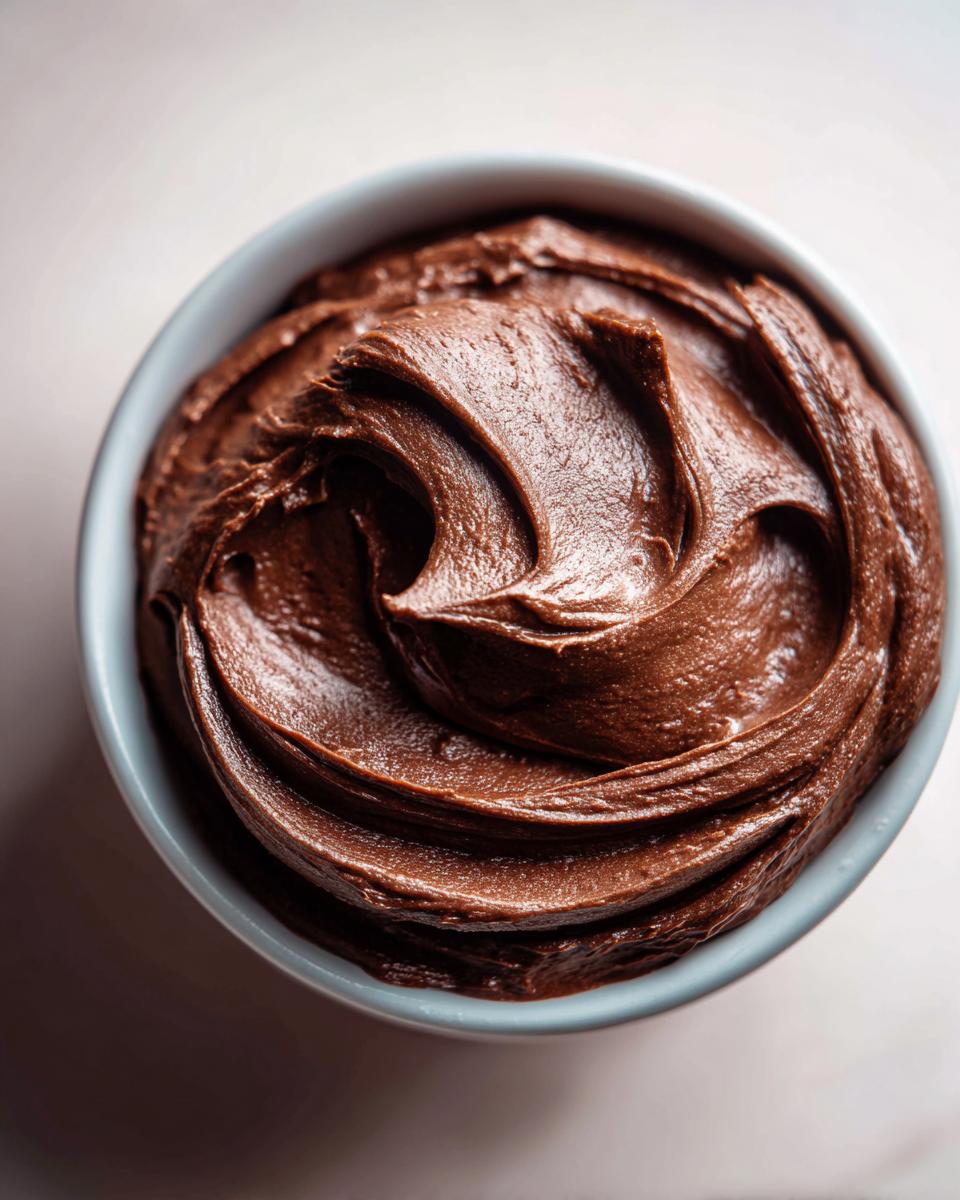 A close-up overhead view of smooth, rich chocolate fudge frosting swirled in a light blue bowl.