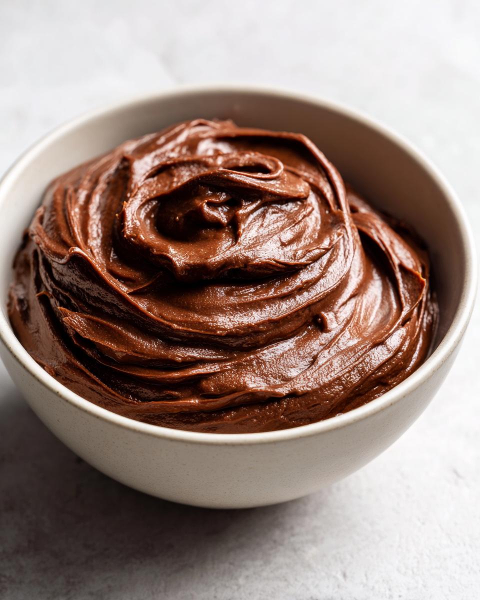 A close-up of smooth, glossy chocolate fudge frosting swirled in a white bowl.