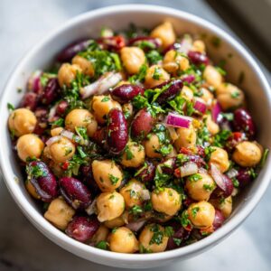 Close-up of a bowl filled with a dense bean salad featuring chickpeas, kidney beans, red onion, and fresh parsley.