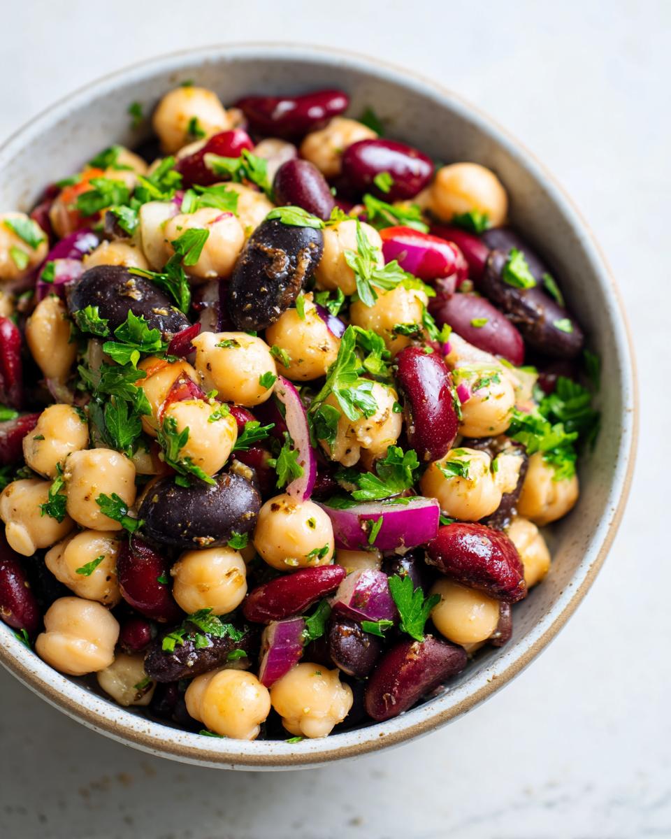 A close-up of a bowl filled with a dense bean salad featuring chickpeas, kidney beans, black beans, and red onion, garnished with fresh parsley.