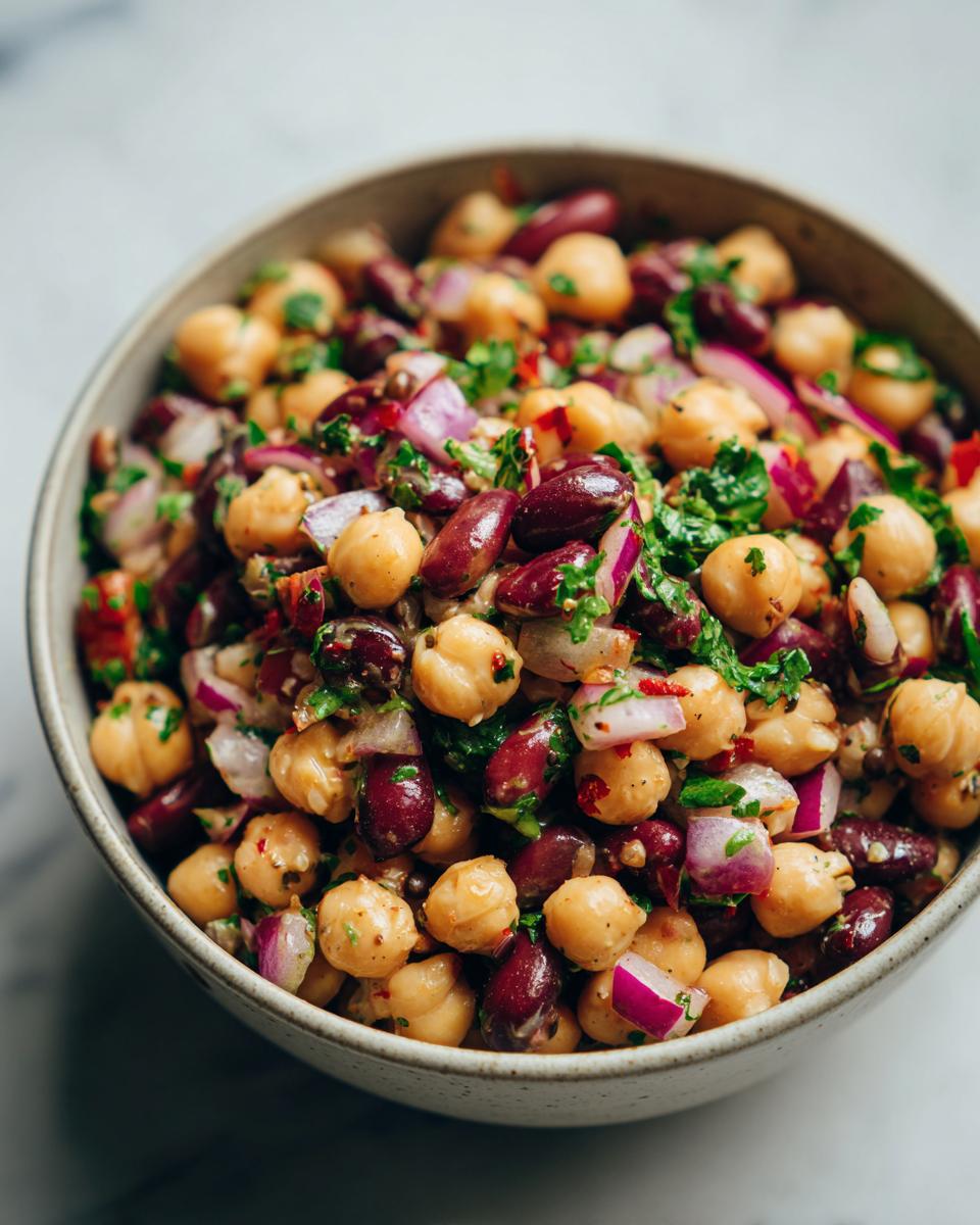 Close-up of a bowl filled with a dense bean salad featuring chickpeas, kidney beans, red onion, and fresh herbs.