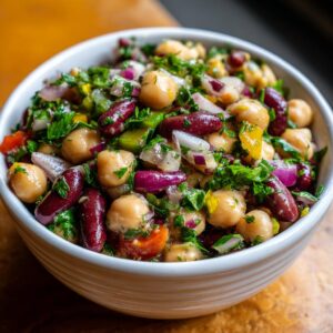 A close-up of a white bowl filled with a vibrant dense bean salad, featuring chickpeas, kidney beans, red onion, and fresh parsley.