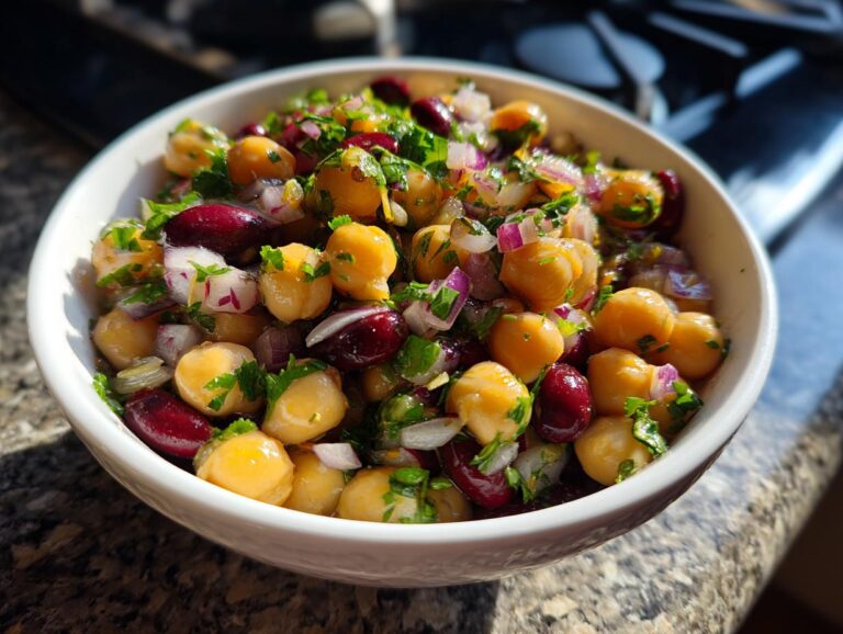 A close-up of a white bowl filled with a dense bean salad, featuring chickpeas, kidney beans, red onion, and fresh parsley.