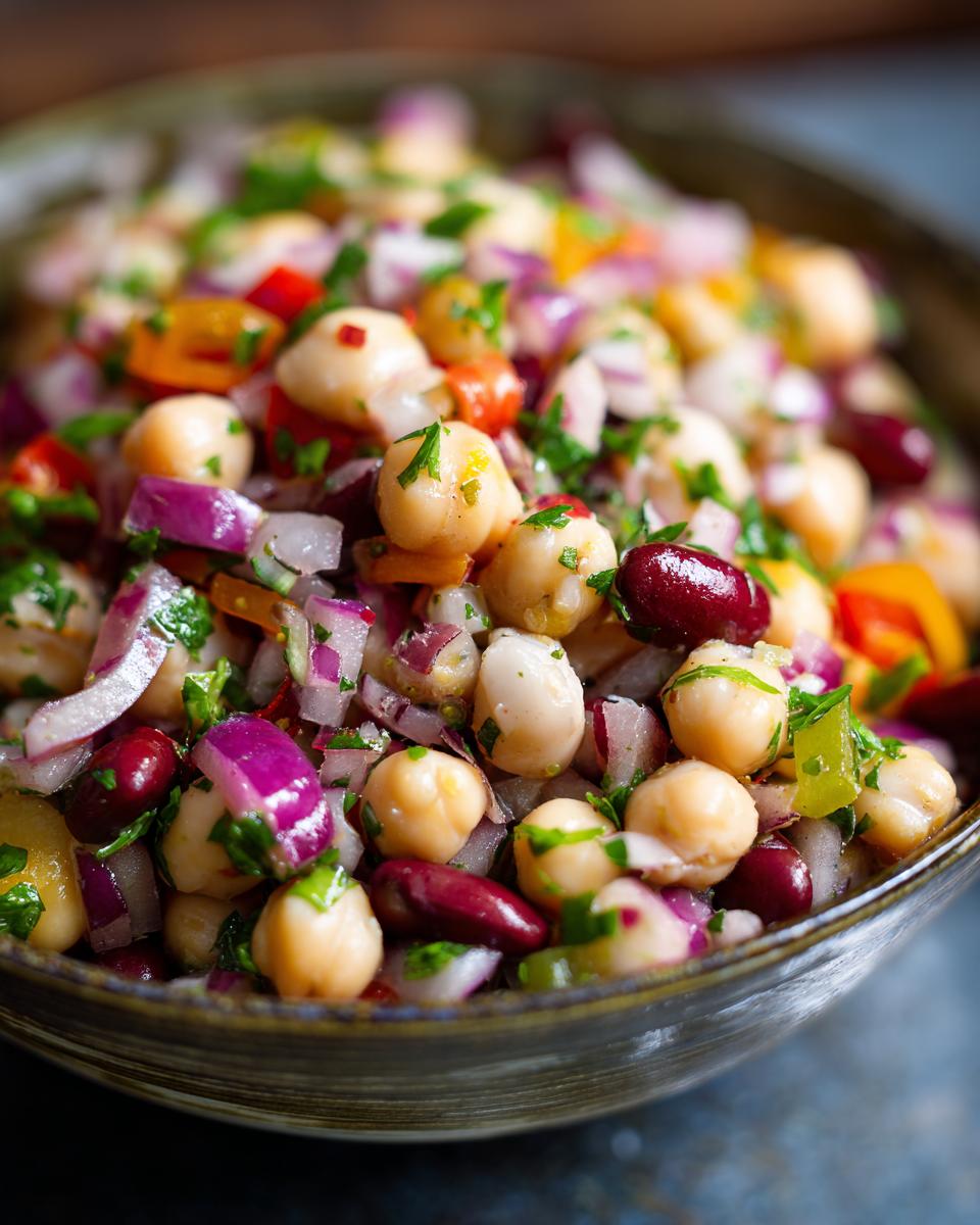 A close-up shot of a vibrant dense bean salad featuring chickpeas, kidney beans, red onion, and bell peppers, garnished with fresh parsley.