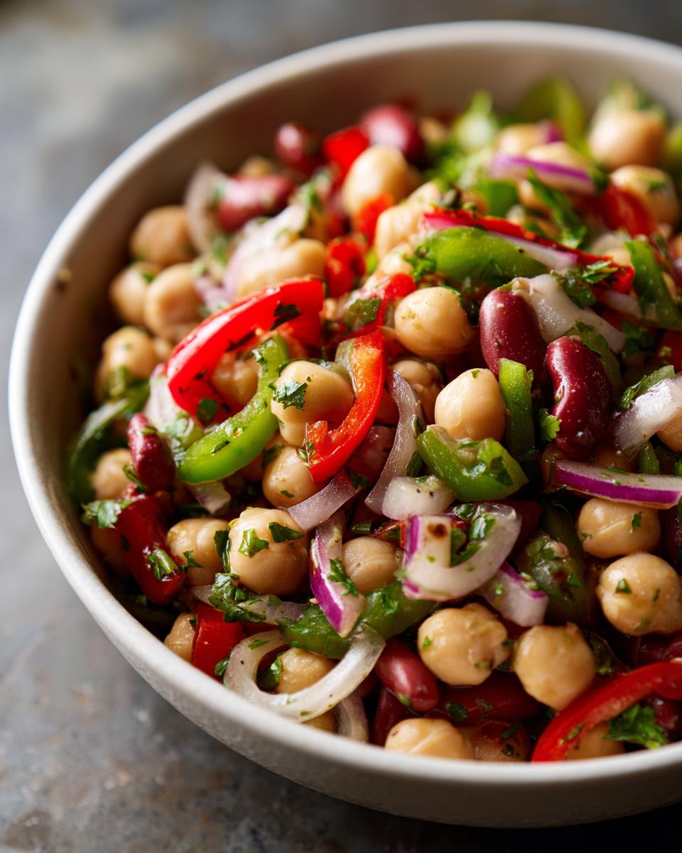 A close-up view of a vibrant dense bean salad featuring chickpeas, kidney beans, red and green bell peppers, and red onion.