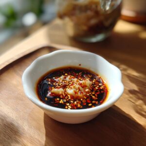 A close-up of a white bowl filled with a rich, dark Dumpling Sauce topped with chili flakes and sesame seeds.