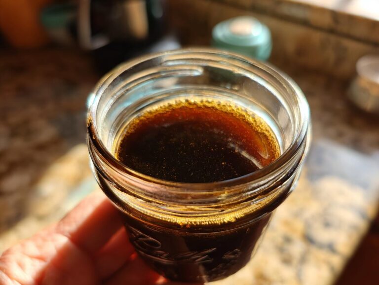 Close-up of a hand holding a small glass jar filled with dark, rich elderberry syrup.