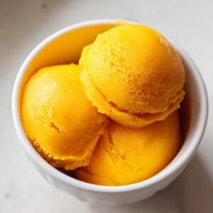 Close-up of three scoops of vibrant orange mango sorbet in a white bowl.