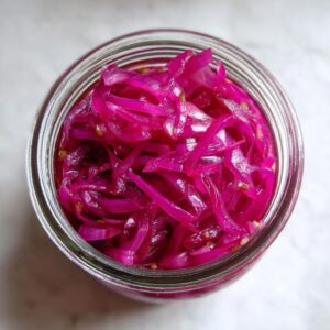 Top-down view of a glass jar filled with bright pink pickled red onions and visible mustard seeds.