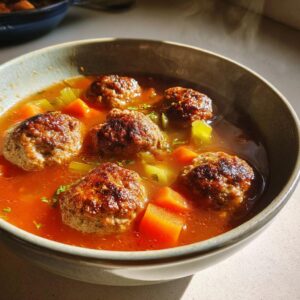 A close-up shot of a bowl of steaming Albondigas soup, featuring savory meatballs and chunks of carrots and celery in a rich broth.