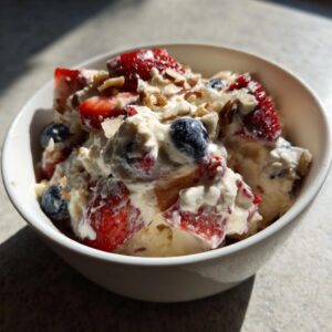 A close-up of a white bowl filled with creamy cheesecake fruit salad, featuring strawberries, blueberries, and chopped nuts.
