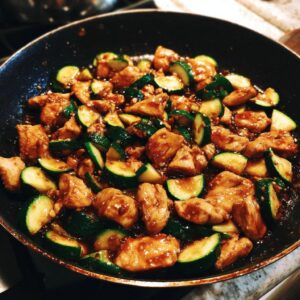 Close-up of chicken zucchini stir fry cooking in a black pan with a glossy sauce.