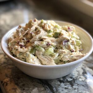 A close-up of a white bowl filled with creamy classic chicken salad, featuring shredded chicken, celery, and red onion.