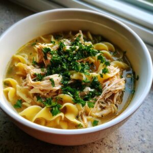 A close-up of a bowl filled with creamy chicken noodle soup, featuring wide egg noodles, shredded chicken, and fresh parsley.