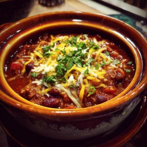 A close-up of a bowl of hearty crockpot chili topped with shredded cheese and fresh chopped herbs.