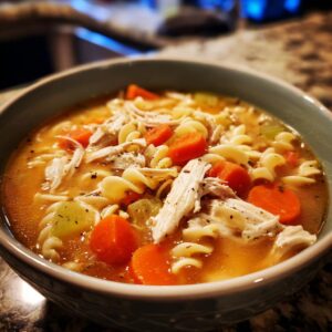 A close-up of a bowl of Italian penicillin soup, featuring shredded chicken, rotini pasta, and bright orange carrot slices in a savory broth.