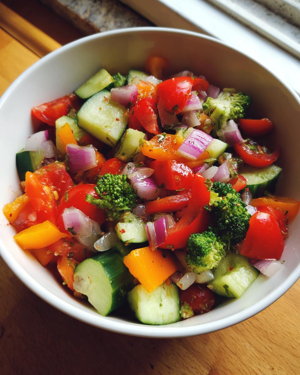 A close-up of a white bowl filled with a vibrant marinated vegetable salad, featuring chopped tomatoes, cucumbers, red onion, bell peppers, and broccoli florets.