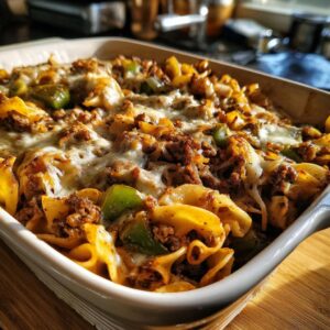 A close-up view of a baked Philly cheesesteak casserole in a white dish, featuring pasta, ground beef, green peppers, and melted cheese.