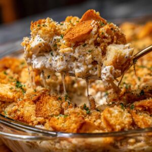 A serving spoon lifts a cheesy, creamy portion of poppy seed chicken casserole, topped with golden breadcrumbs and parsley.