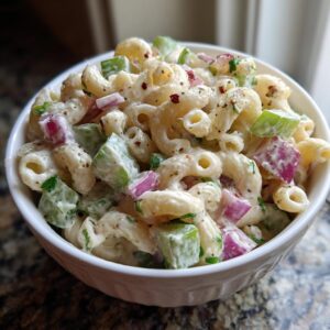 A close-up of creamy Macaroni Salad in a white bowl, featuring elbow macaroni, diced celery, red onion, and herbs.