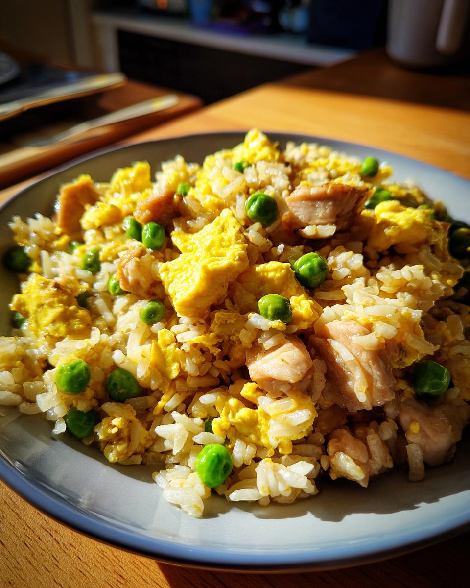 A close-up of a bowl of chicken fried rice, featuring fluffy rice, scrambled egg, tender chicken pieces, and bright green peas.