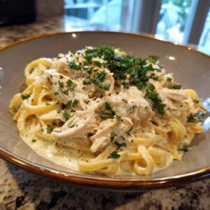 A close-up of a bowl of creamy crockpot chicken alfredo pasta, topped with shredded chicken and fresh parsley.