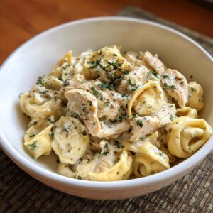 A close-up of a white bowl filled with creamy crockpot chicken alfredo tortellini, garnished with fresh herbs.