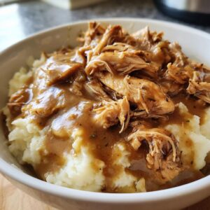 A bowl of creamy mashed potatoes topped with shredded crockpot chicken and gravy, seasoned with herbs.