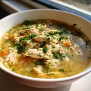 A close-up of a bowl of lemon chicken orzo soup, filled with shredded chicken, orzo pasta, carrots, and fresh herbs.