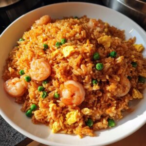 A close-up, appetizing shot of homemade shrimp fried rice with pink shrimp, green peas, and scrambled egg pieces served in a white bowl.