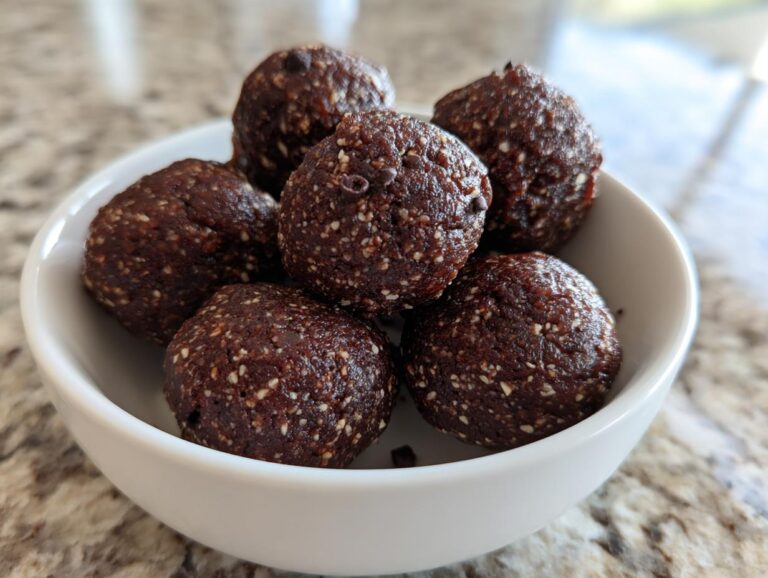 A close-up of several rich, dark chocolate protein balls speckled with white seeds, served in a small white bowl.