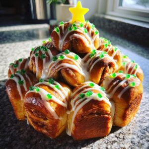 A stack of baked cinnamon rolls arranged in a Christmas tree shape, topped with white icing and green sprinkles, featuring a yellow star.