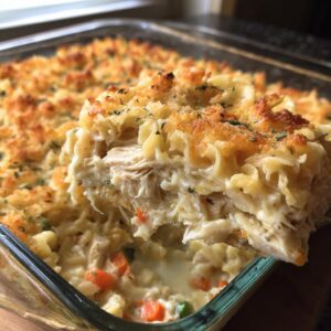 A serving spoon lifts a creamy slice of chicken noodle casserole, showing shredded chicken, noodles, and vegetables under a golden, crunchy topping.
