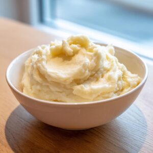 A white bowl filled high with fluffy, creamy mashed potatoes sitting on a wooden table near a window.