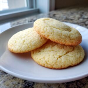 Three freshly baked, slightly golden sugar cookies piled on a white plate, coated in sparkling sugar.