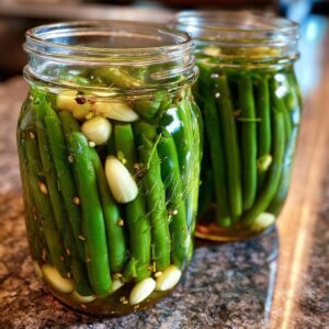 Two glass jars filled with bright green dilly beans, garlic cloves, and pickling spices ready for canning.