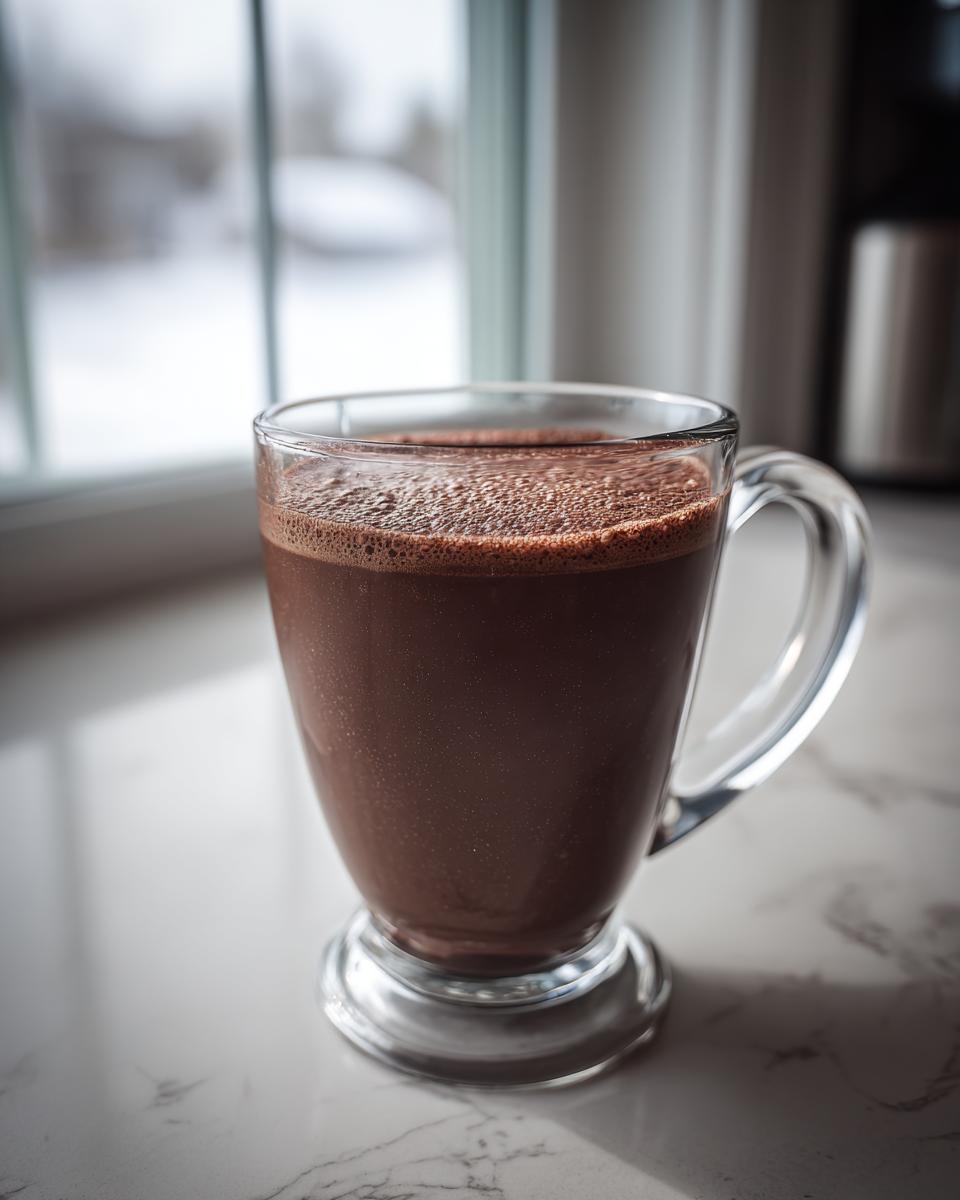 A clear glass mug filled with rich, frothy hot cocoa mix sitting on a marble counter near a window showing a snowy scene.