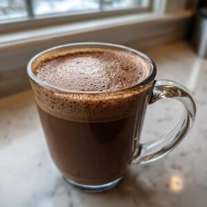 Close-up of a clear glass mug filled with rich, dark hot cocoa mix topped with thick foam.