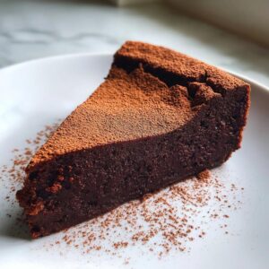 A rich, fudgy slice of flourless chocolate torte dusted heavily with cocoa powder on a white plate.