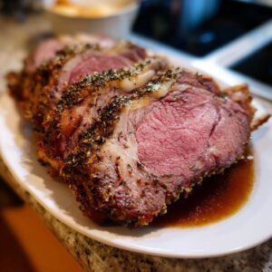 Close-up of thick, medium-rare slices of herb-crusted prime rib resting in its juices on a white platter.