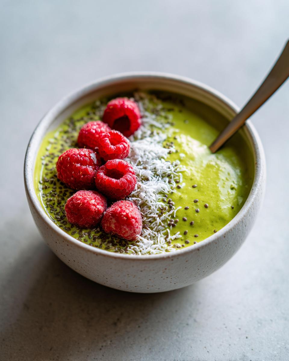 A bowl of vibrant green matcha chia pudding topped with fresh raspberries, shredded coconut, and chia seeds.