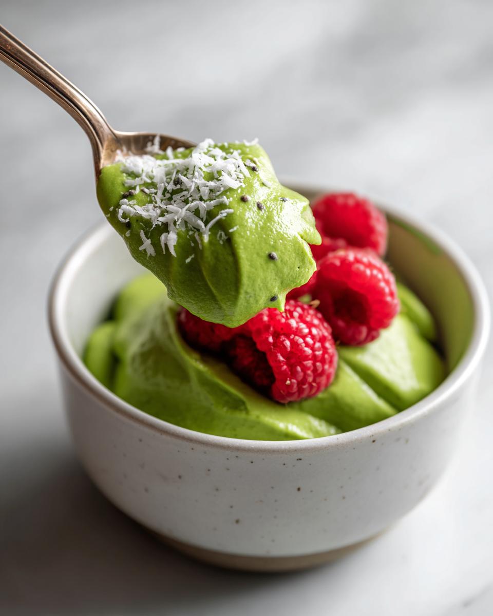 A spoonful of vibrant green matcha chia pudding topped with shredded coconut being lifted from a bowl garnished with fresh raspberries.