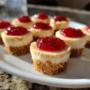 Close-up of several delicious no-bake cheesecake bites topped with bright red strawberry jam on a white plate.