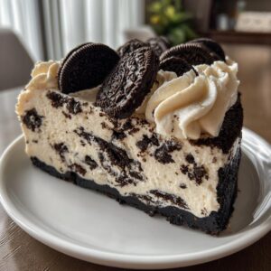 Close-up of a rich slice of no bake Oreo cheesecake with a dark crust, creamy filling mixed with cookie pieces, and topped with whipped cream and whole Oreos.