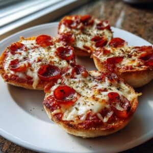Four perfectly baked pepperoni english muffin pizza halves topped with melted cheese and herbs on a white plate.