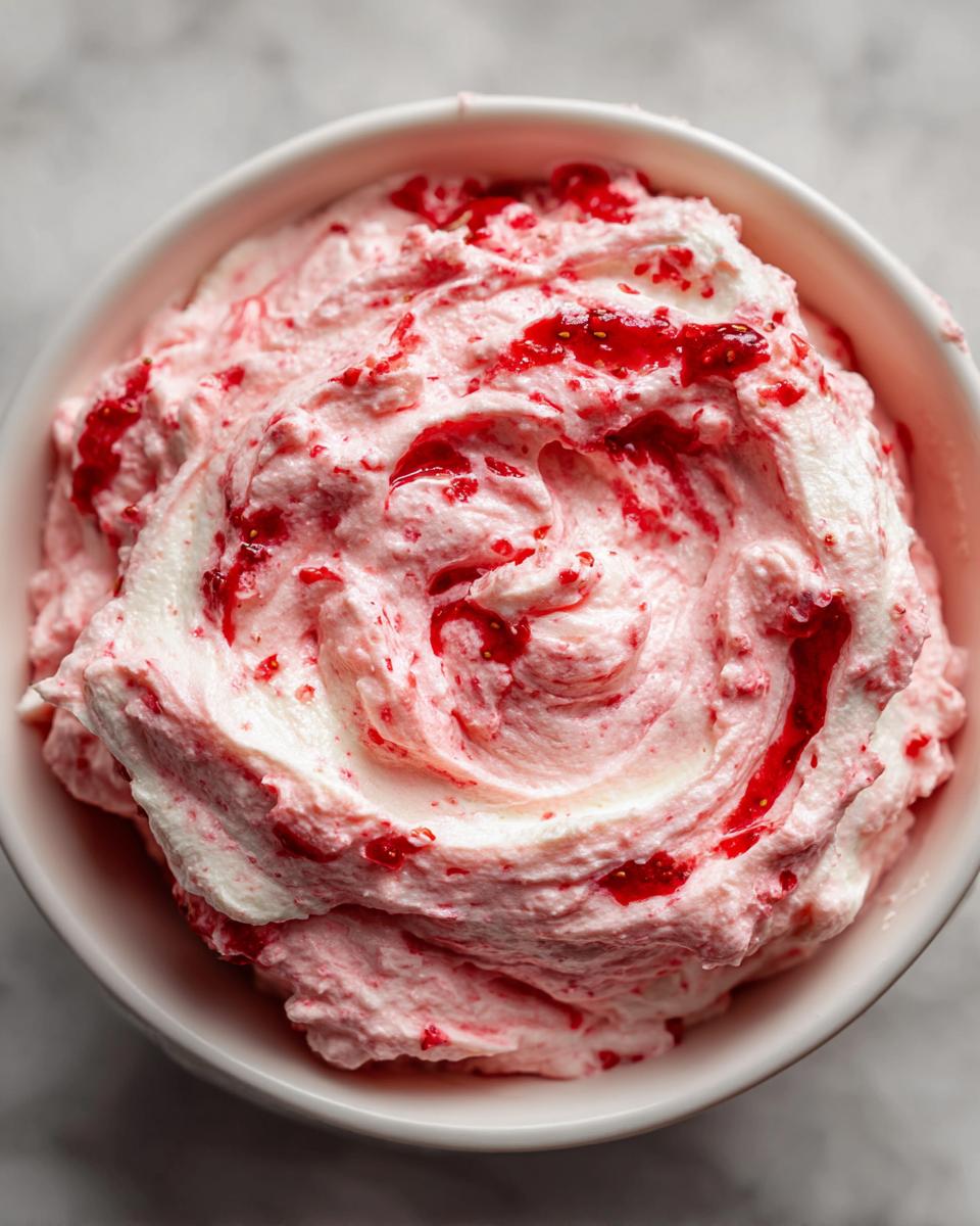 Close-up overhead shot of fluffy strawberry cheesecake dip swirled with bright red strawberry preserves in a white bowl.