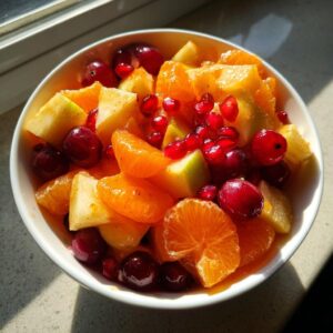 Close-up of a bowl filled with a vibrant winter fruit salad featuring mandarin segments, cranberries, pomegranate seeds, and apple chunks.